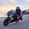 2025 Yamaha YZF-R1M sportbike on track. Rider in full gear, Arai helmet. Black and blue color scheme. High-performance motorcycle.