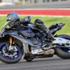 2025 Yamaha YZF-R1M motorcycle on the track. Rider in full gear leans into a turn. Black and blue sportbike.