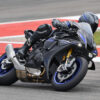2025 Yamaha YZF-R1M action shot. Rider in full gear leans into a turn on the track. Black and blue sportbike.