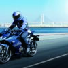 A person wearing a helmet rides a blue SUZUKI GIXXER SF 250 2024 motorcycle along a scenic coastal highway with a bridge visible in the background.