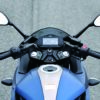 The SUZUKI GIXXER SF 250 2024's handlebar showcases a sleek digital display, rearview mirrors, and easy-grip controls on both sides, set against the backdrop of the road speeding by.