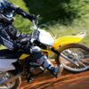 In black and blue gear, a skilled rider maneuvers a yellow SUZUKI DR-Z125L LARGE WHEEL dirt bike through a tough track, gracefully leaning into a curve with lush greenery in the backdrop.