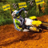 Dressed in yellow and white, the rider skillfully leans into a turn on a SUZUKI RM-Z450, expertly navigating the dirt track. In the background, Suzuki banners wave proudly, enhancing the thrilling atmosphere.