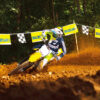 A motocross rider expertly maneuvers a dirt track on a yellow SUZUKI RM-Z450, causing dust to fly. "Suzuki" banners and checkered flags wave in the background among trees.