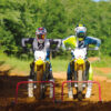 Two motocross riders, equipped with helmets and gear, rev up their engines at the starting gate on a dirt track. A reliable SUZUKI RM-Z450 is poised for action, with trees framing the backdrop.