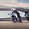 Two motorcyclists in protective gear ride CFMOTO 650 GT ABS bikes on a road beside a large body of water, with mountains in the background.