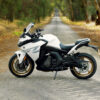A sleek, white CFMOTO 650 GT ABS sport motorcycle is parked on a deserted, tree-lined road, embraced by vibrant foliage on either side.