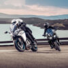 Two motorcyclists wearing helmets navigate a winding road by water, framed by hills, demonstrating the agility of their CFMOTO 650 GT ABS bikes.