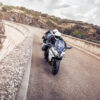 Two motorcyclists wearing helmets ride their CFMOTO 650 GT ABS bikes on a winding road near a dam wall and rocky landscape under a partly cloudy sky.