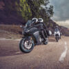 Two motorcyclists in protective gear ride CFMOTO 650 GT ABS bikes along a winding mountain road, with trees on one side and a stunning water view on the other.