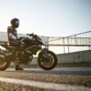 Dressed in full riding gear, a person is seated on a sleek black CFMOTO 800NK SPORT motorcycle parked on a paved road, with a bridge and fence as the backdrop under a clear sky.