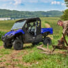 Blue 2024 Yamaha Viking III UTV. Man using chainsaw. Utility vehicle in field.