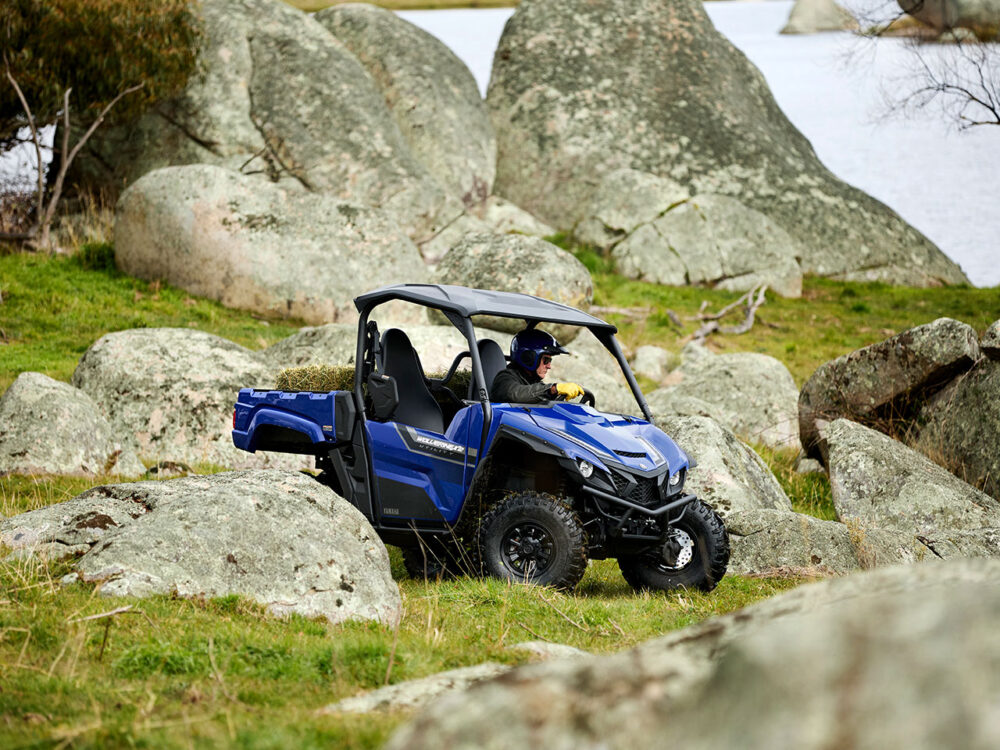 A person navigates a YAMAHA WOLVERINE X2 UTILITY, colored blue, across rocky grassland near water with large boulders scattered around the terrain.