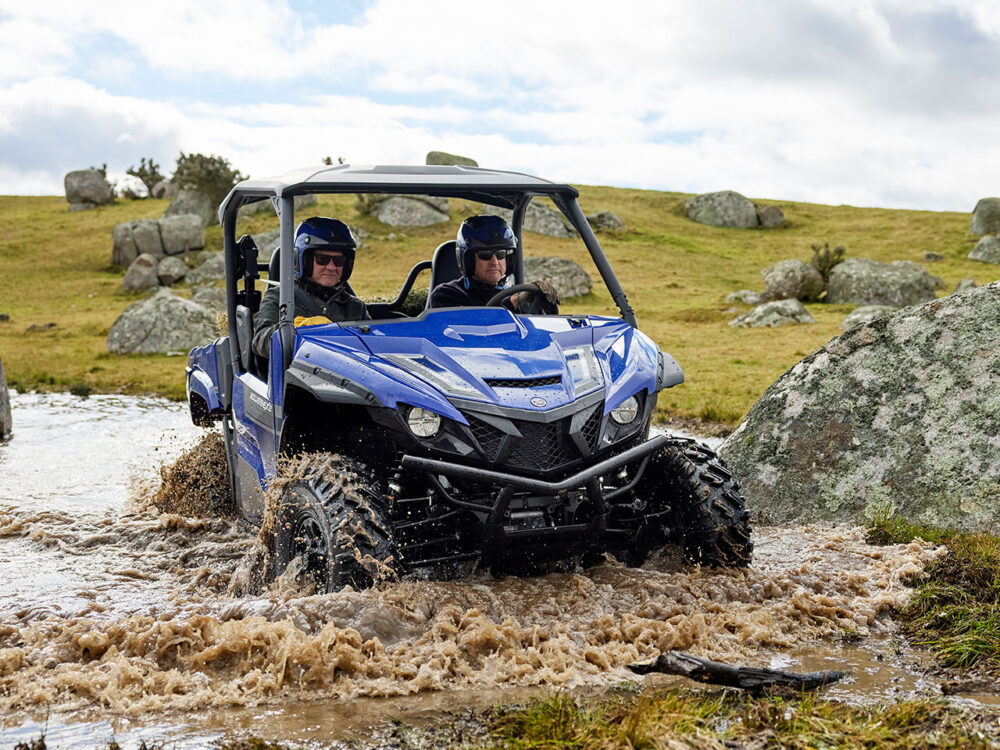 Two people drive a YAMAHA WOLVERINE X2 UTILITY, a blue off-road vehicle, through muddy water on a grassy and rocky landscape.