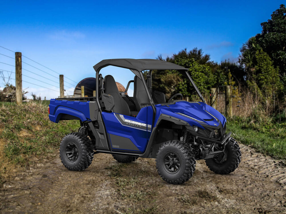 A YAMAHA WOLVERINE X2 UTILITY, an off-road vehicle in blue, is parked on a dirt path surrounded by grass and trees under a clear blue sky.