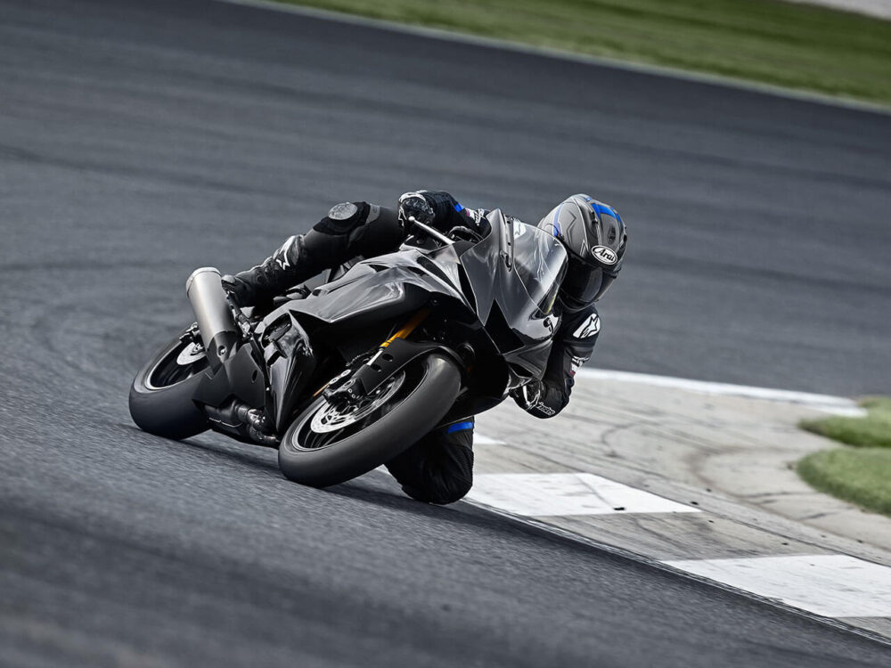 A motorcyclist in full gear leans into a turn on a racetrack, riding a sleek, dark-colored YAMAHA YZF-R6 RACE 2023. The track is bordered by grass.