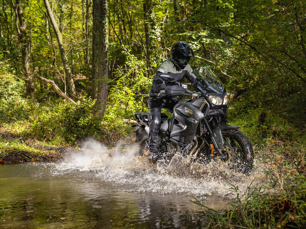 A YAMAHA SUPER TENERE rider navigates a shallow water crossing in a dense forest, causing splashes.