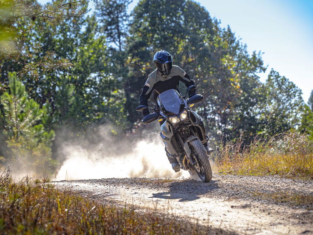 A rider on a YAMAHA SUPER TENERE navigates a dirt trail, kicking up dust amidst trees and grass.