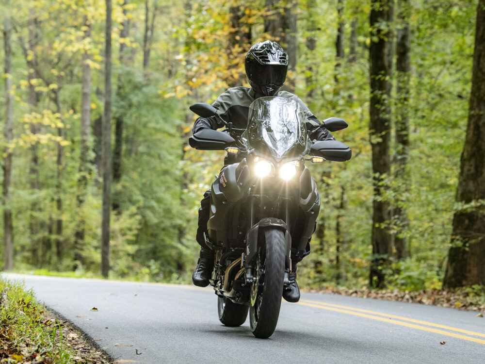 A person rides a YAMAHA SUPER TENERE on a tree-lined road, surrounded by greenery, during the day.