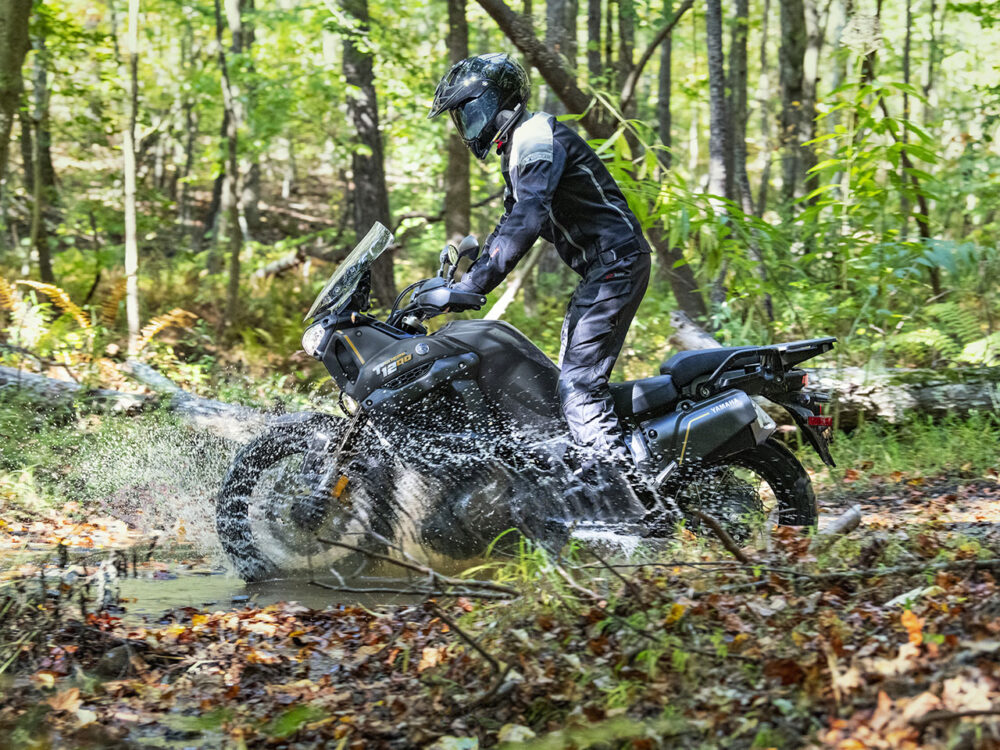 Someone rides a YAMAHA SUPER TENERE through a muddy forest trail, creating splashes of water and mud.