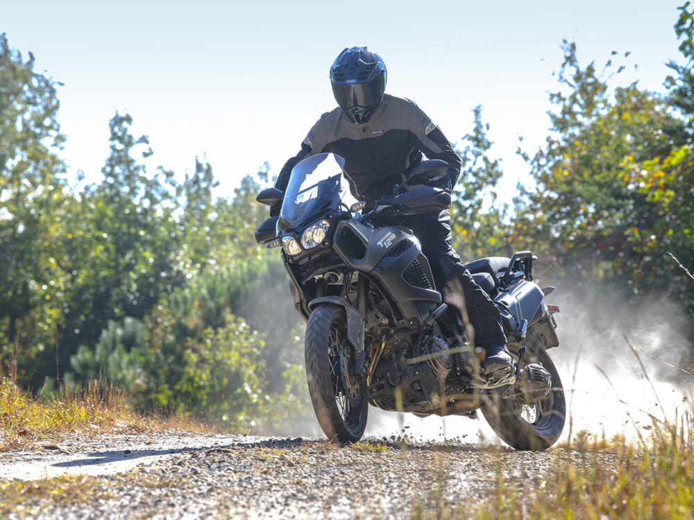 A motorcyclist rides a YAMAHA SUPER TENERE on a gravel path in a forest, creating dust trails. The rider is wearing protective gear and a helmet.