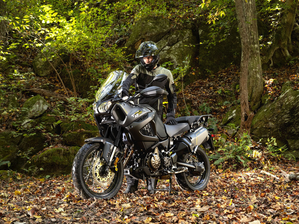 A rider in full gear stands beside a YAMAHA SUPER TENERE, an adventure motorcycle, amidst a leaf-covered forest floor.