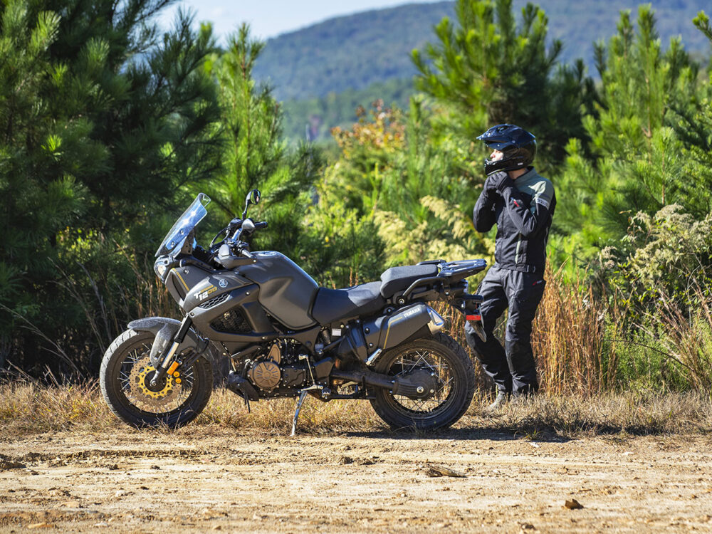 A motorcyclist adjusts their helmet near a parked YAMAHA SUPER TENERE on a dirt path, surrounded by trees and hills.