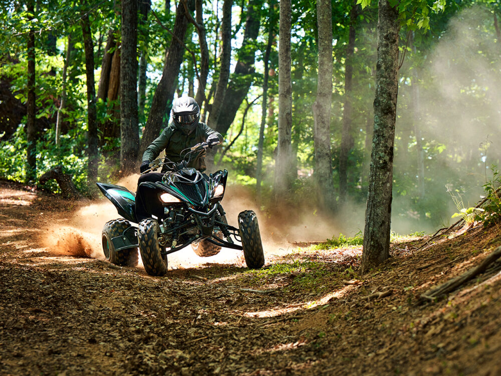 A rider on the 2024 YAMAHA YFM700R powers through a forest trail, surrounded by lush greenery and kicking up dust.