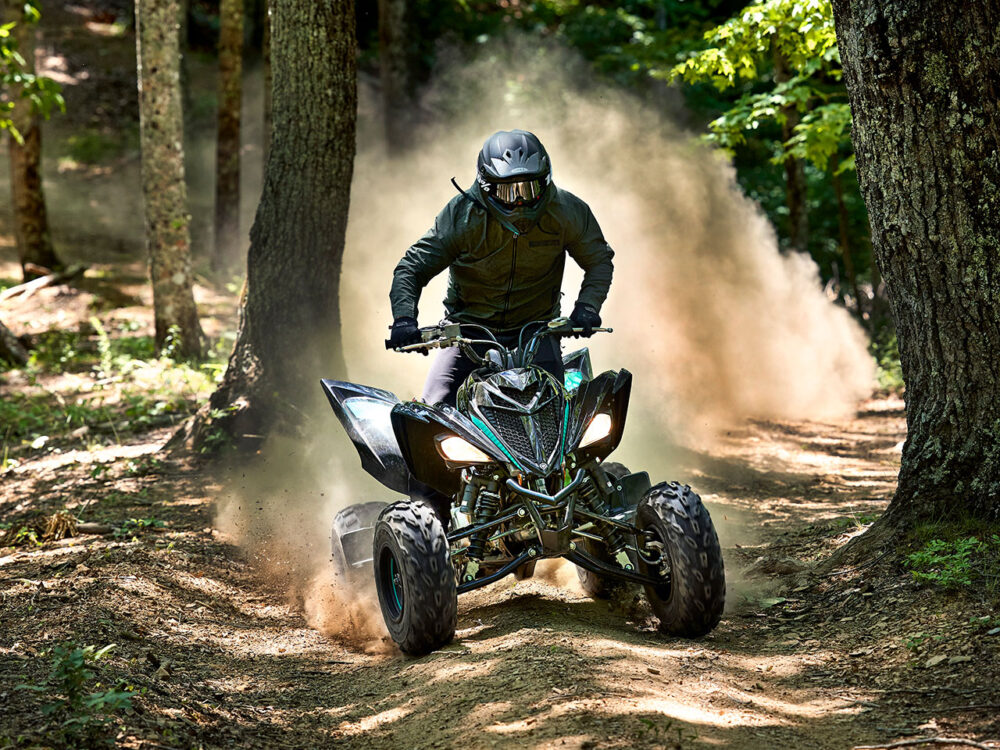 A person rides the 2024 YAMAHA YFM700R through a forest trail, surrounded by trees, kicking up dust.