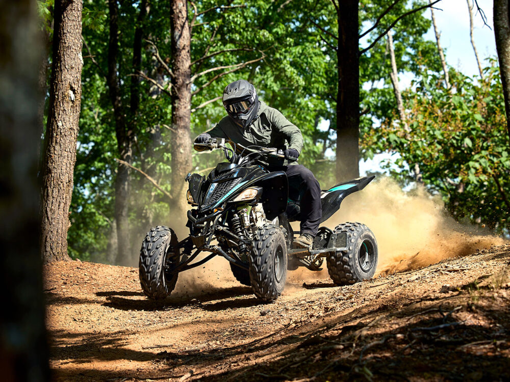 A person wearing a helmet rides the YAMAHA YFM700R - 2024 ATV through a forest dirt trail, kicking up dust.