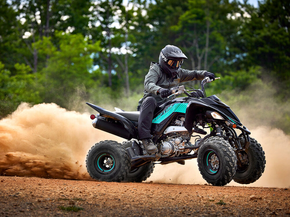 A rider on a YAMAHA YFM700R - 2024 ATV speeds along a dirt path, kicking up dust amidst trees and greenery.