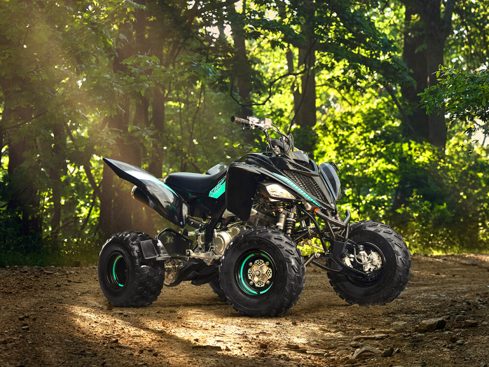 A YAMAHA YFM700R - 2024 ATV with a sleek black design is parked on a dirt path in a sunlit forest.