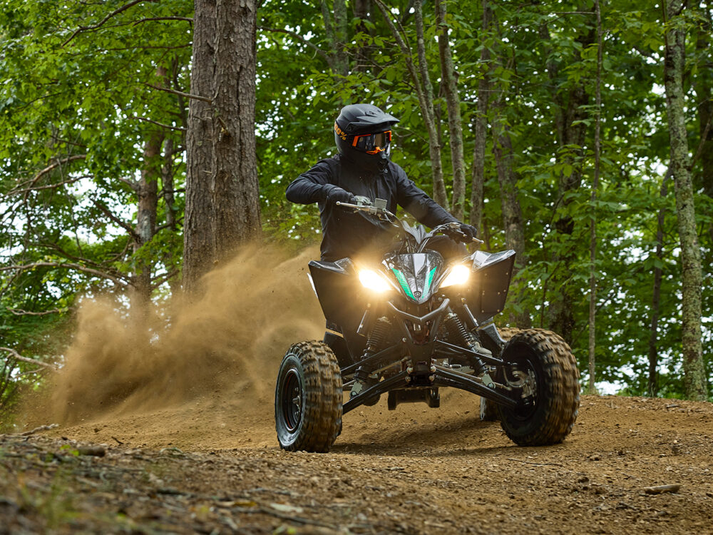 A person rides a YAMAHA YFZ450R 2024 ATV through a forest trail, kicking up dirt with trees in the background.