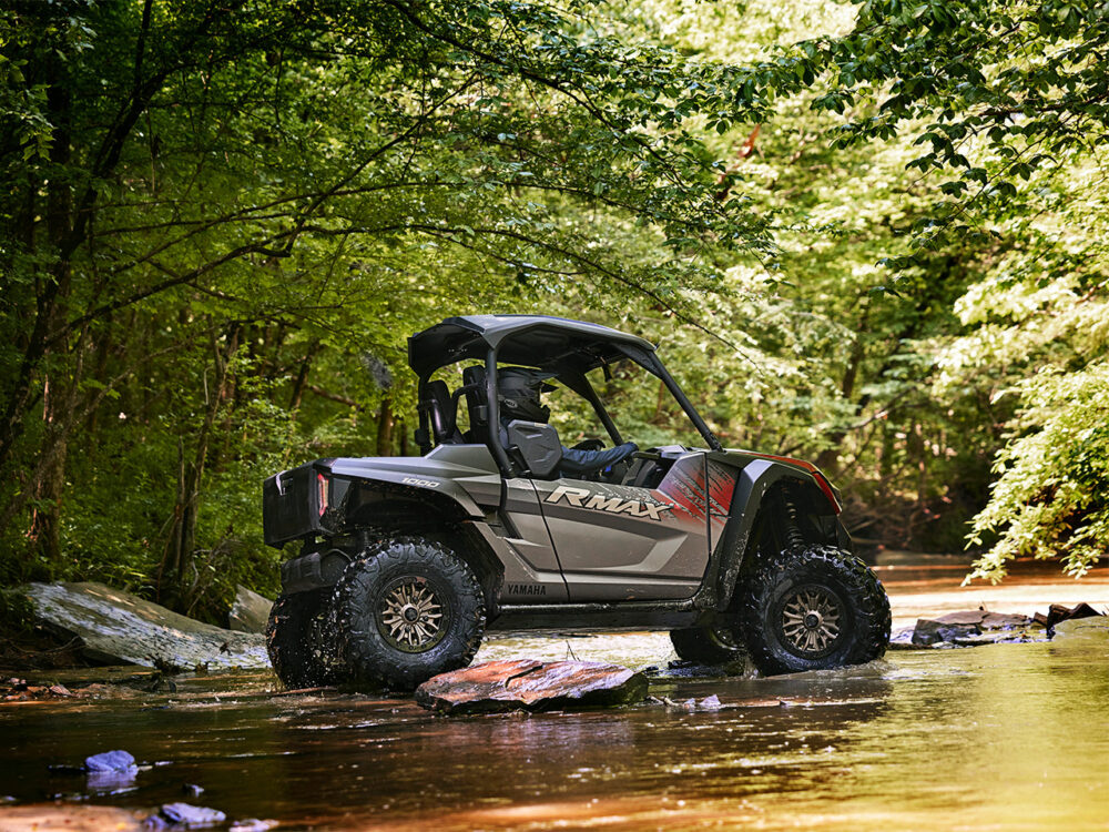 A YAMAHA RMAX2 1000 XT-R off-road vehicle is positioned on large rocks in a shallow, wooded creek.