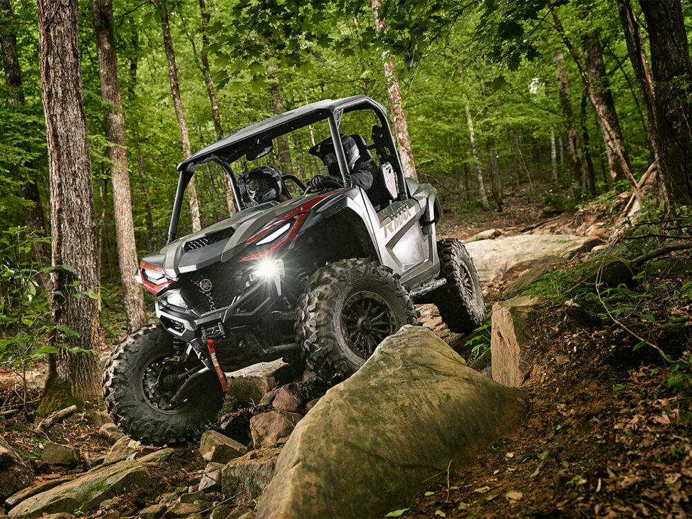 The YAMAHA RMAX2 1000 XT-R skillfully navigates a rocky forest path, surrounded by tall trees and dense greenery.