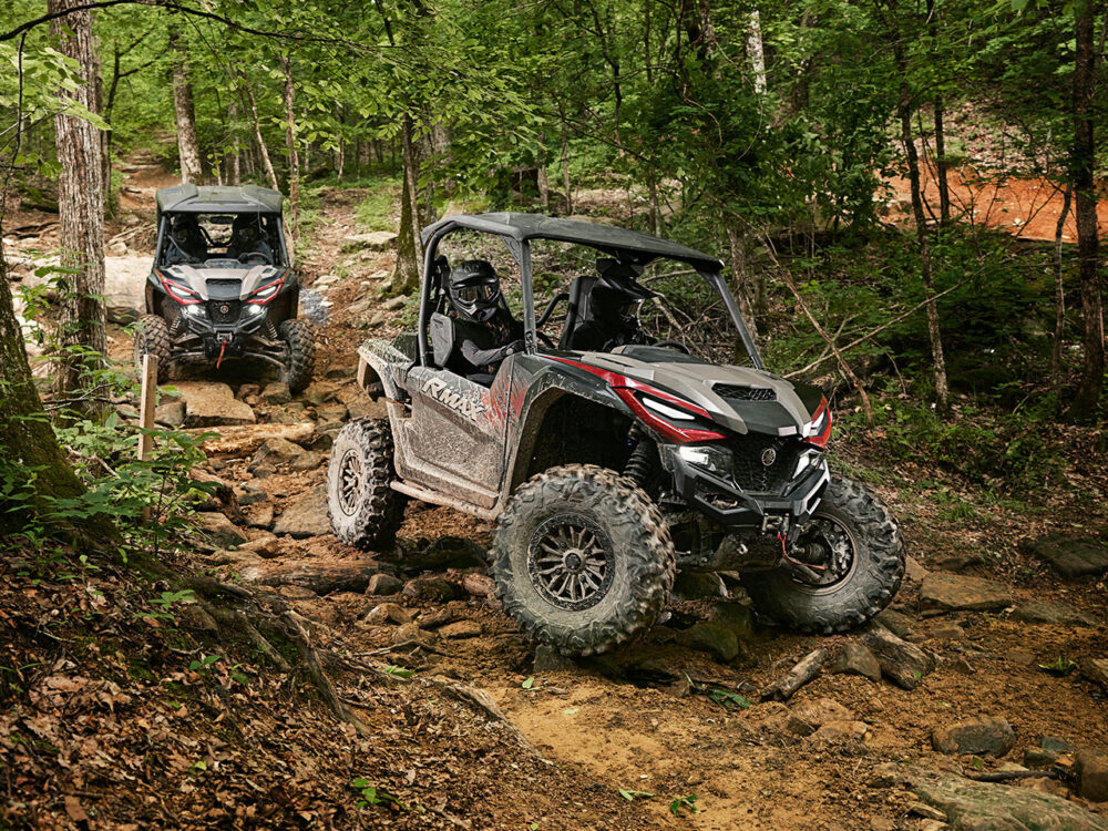 A YAMAHA RMAX2 1000 XT-R navigates a rocky forest trail, surrounded by dense green foliage.