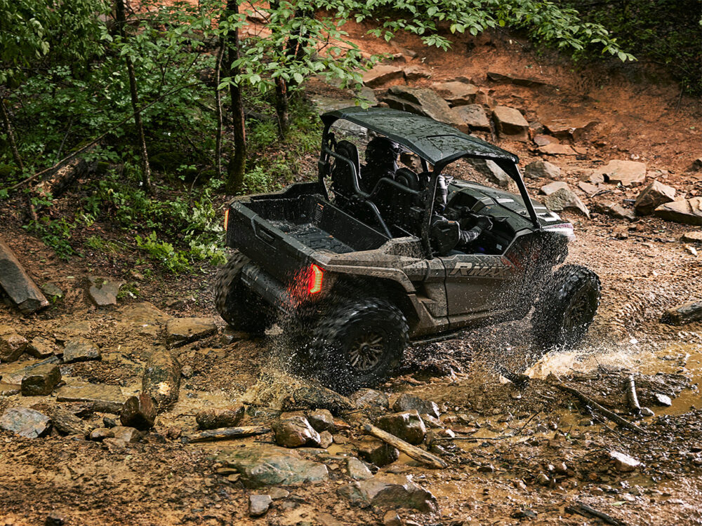 The YAMAHA RMAX2 1000 XT-R powers through a muddy forest trail, splashing water and maneuvering expertly among trees and rocks.