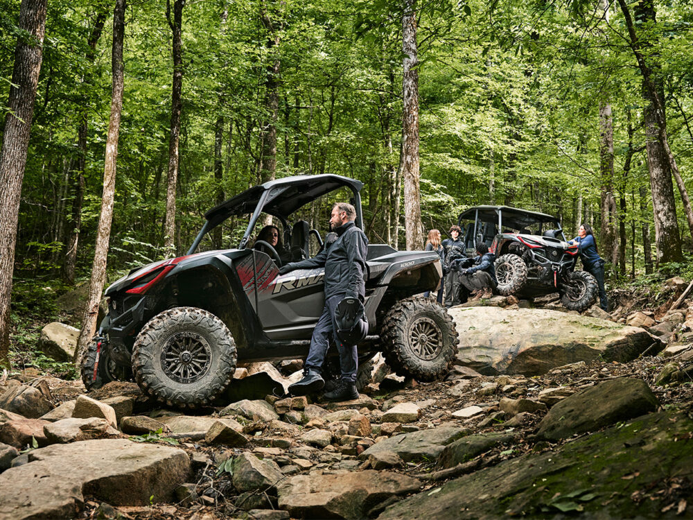 Two YAMAHA RMAX2 1000 XT-R off-road vehicles, each carrying several people, navigate a rocky forest trail surrounded by dense green trees.