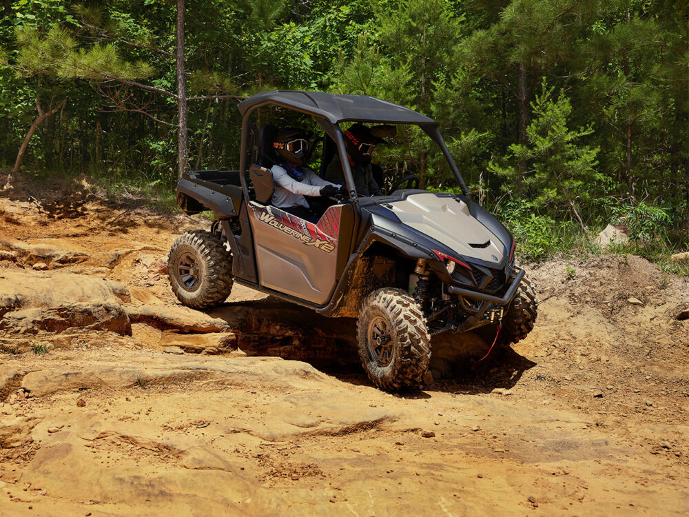 Two people drive a YAMAHA WOLVERINE X2 XT-R over a rocky dirt trail surrounded by trees.