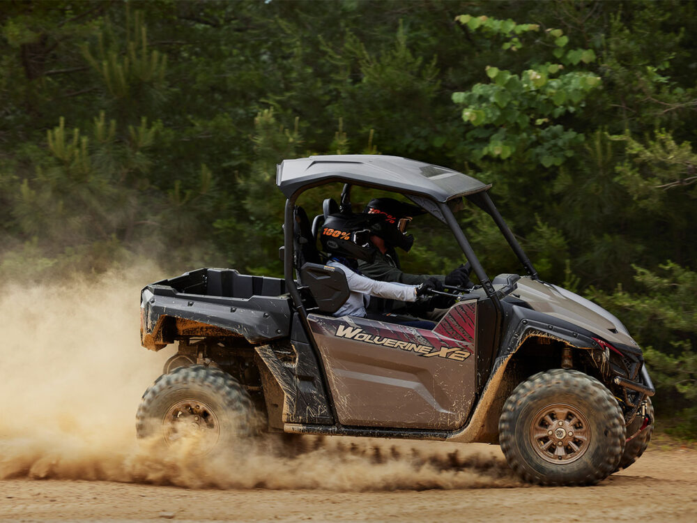 A person drives a "Yamaha Wolverine X2 XT-R" off-road vehicle on a dirt path, kicking up dust, with trees in the background.