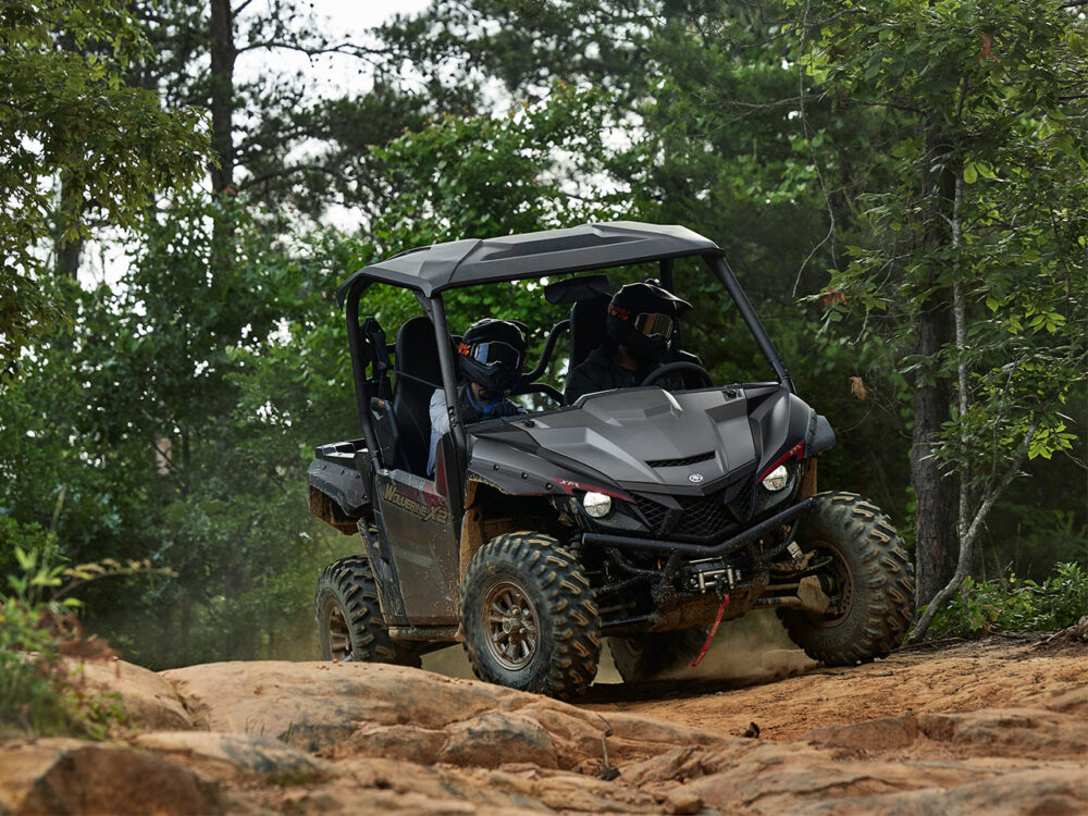 Two people navigate rocky terrain in a YAMAHA WOLVERINE X2 XT-R, surrounded by trees and lush greenery.