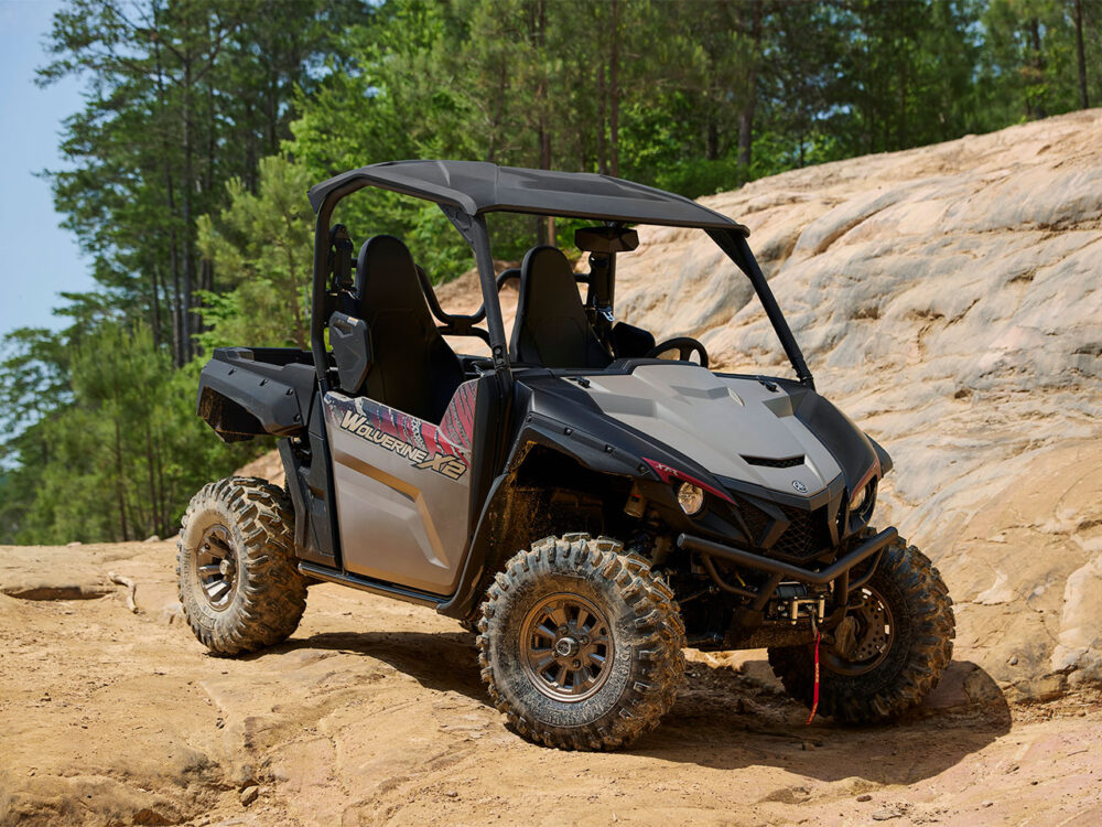 A YAMAHA WOLVERINE X2 XT-R, equipped with a roll cage and rugged tires, navigates rocky terrain surrounded by trees.