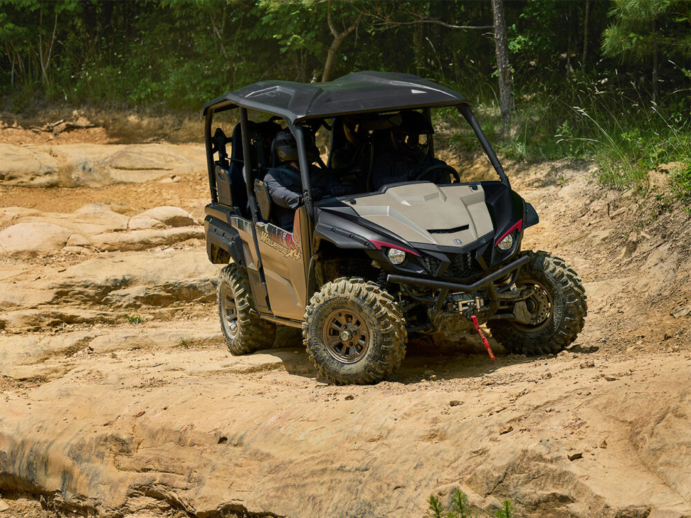 The YAMAHA WOLVERINE X4 XT-R, a rugged off-road vehicle with a roll cage, traverses rocky, uneven terrain surrounded by sparse vegetation and trees.