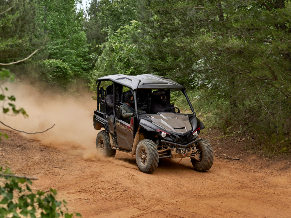 Two passengers ride the YAMAHA WOLVERINE X4 XT-R along a dusty dirt path in a forested area.