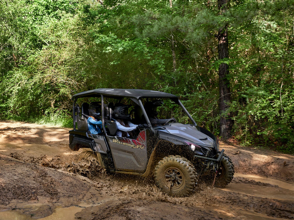 A YAMAHA WOLVERINE X4 XT-R navigates muddy terrain in a forested area with passengers.