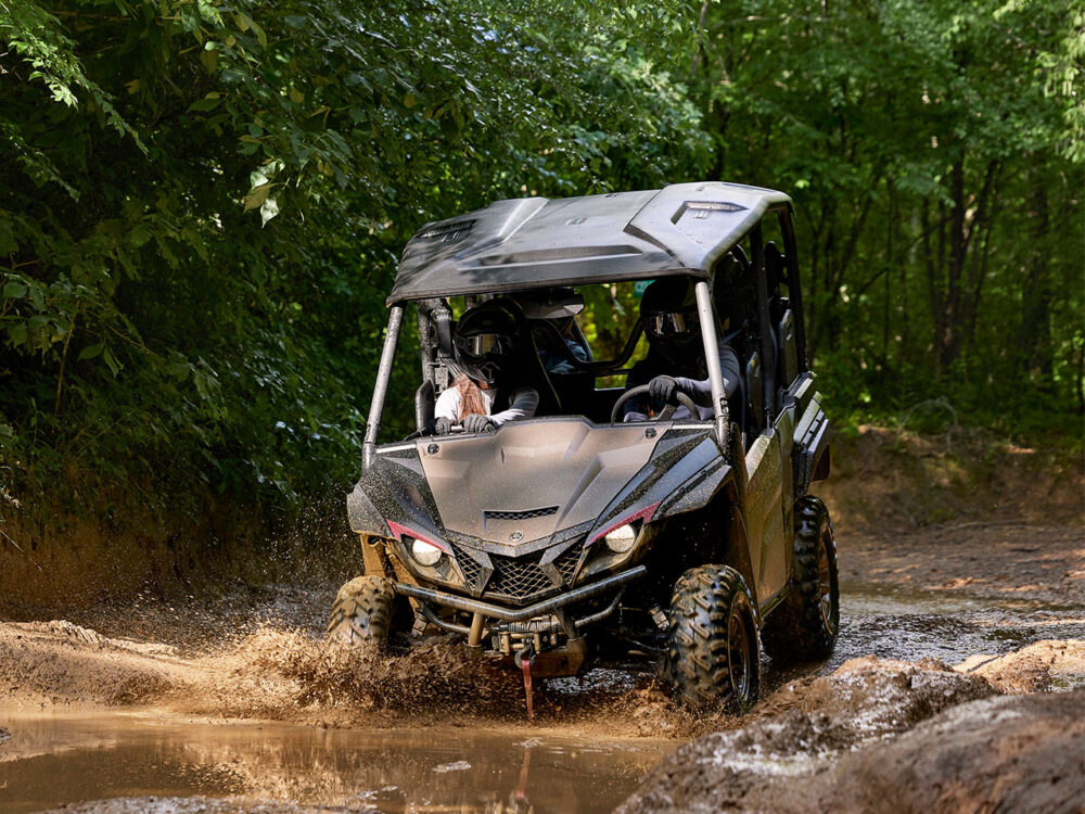 The YAMAHA WOLVERINE X4 XT-R powers through a muddy forest trail, kicking up splashes of water and mud.