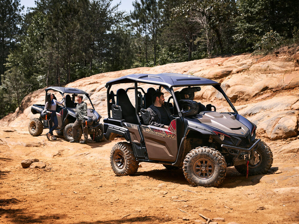 Two YAMAHA WOLVERINE X4 XT-R off-road vehicles with passengers navigate a rocky dirt trail surrounded by trees.