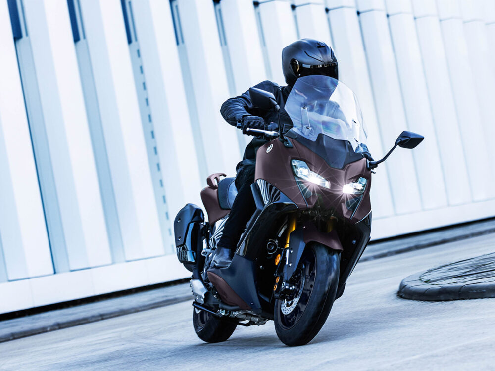 A rider in black helmet and gear navigates a curved road on a YAMAHA TMAX 560 - 2024, adjacent to a modern white building.