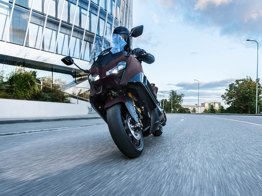 A rider on a YAMAHA TMAX 560 - 2024, with a helmet on, is traveling down an empty city road beside a sleek glass building.