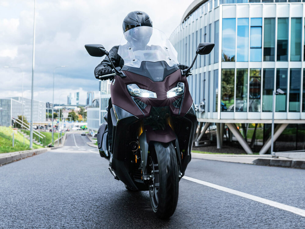 A motorcyclist in black gear rides a YAMAHA TMAX 560 - 2024 scooter on an urban road, surrounded by buildings and under a cloudy sky.